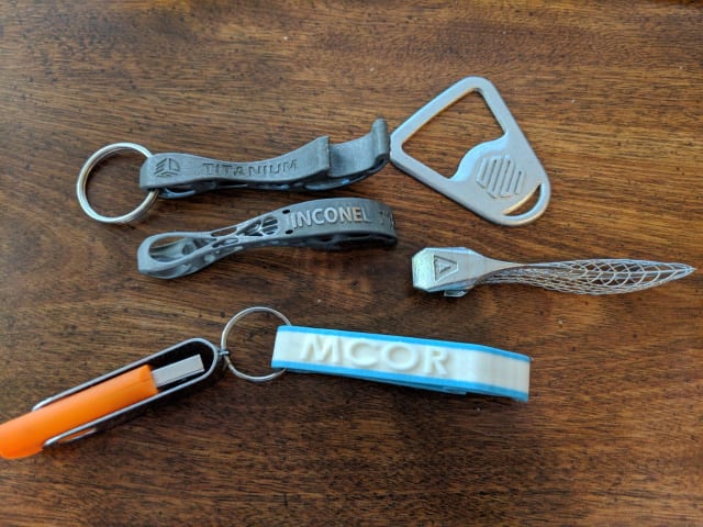 [3D printed bottle openers circa 2018, with models from 3D Systems (printed in titanium and Inconel), Markforged, Velo3D, and &ndash; 3D printed in paper with an attached media kit USB &ndash; MCor // Image Source: Author]